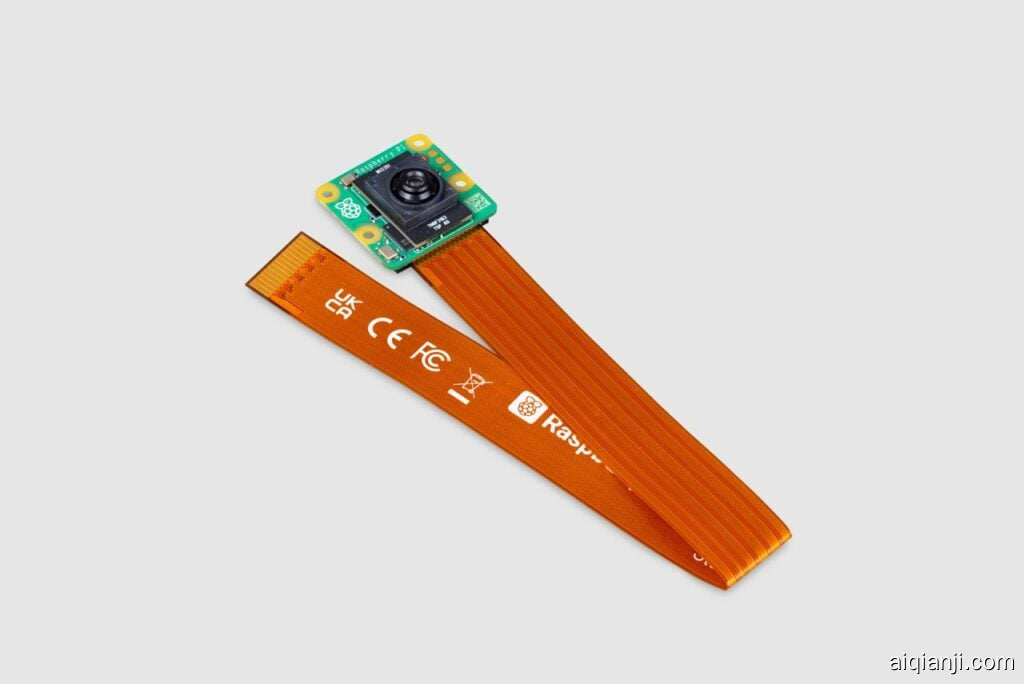 这张图片展示了一个连接在长而扁平的橙色软排线上的树莓派 AI 摄像头模块。摄像头模块小巧且方形，呈绿色，并在中心有一个黑色镜头。每个角落都有黄色的安装孔。附着在模块上的橙色软排线上印有各种法规标志和标识，如 "UKCA"、"CE"、"FCC" 以及树莓派的标志。该电缆看起来非常灵活，专门设计用于连接摄像头和树莓派。摄像头放置在一个浅灰色背景上。